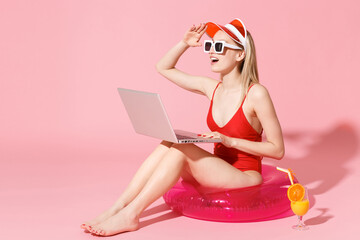 Wall Mural - Young woman in red one-piece swimsuit cap sunglasses isolated on pink background. People summer vacation rest concept. Sit in swim inflatable ring work on laptop pc computer looking far away distance.