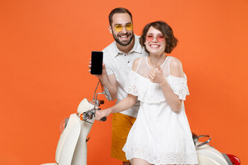 Wall Mural - Smiling couple friends guy girl in summer clothes glasses sit on moped isolated on orange background. Driving motorbike transportation concept. Hold mobile phone with empty screen, showing thumb up.