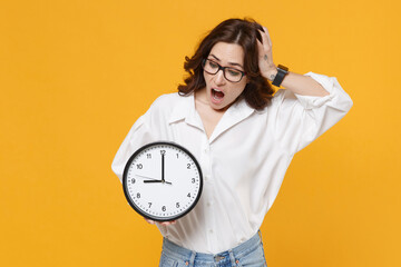 Wall Mural - Shocked young brunette business woman in white shirt glasses isolated on yellow wall background studio. Achievement career wealth business concept. Mock up copy space. Hold clock, put hand on head.