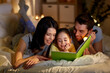© Syda Productions - people and family concept - happy mother, father and little daughter reading book with torch light in bed at night at home
