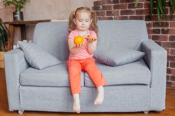 beautiful little girl holding pizza and an orange in her hands. the concept of choosing a healthy di
