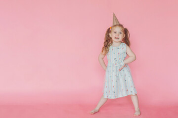 A little cute happy girl in birthday cap  grimaces with happy face on a pink background. Copy space