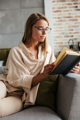 Poster - Image of young concentrated woman reading book while sitting on couch