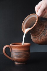 Wall Mural - Milk flowing from a clay jug in a man’s hand onto a clay mug on a black background