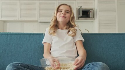 Sticker - A smiling little girl is eating popcorn while watching TV at home in the living room