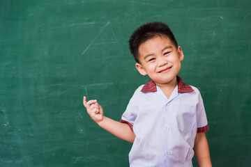 Back to School. Happy Asian funny cute little child boy from kindergarten in student uniform smiling point finger to side away space on green school blackboard, First time to school education concept