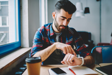 Cheerful hipster guy dressed in casual wear managing time on wristwatch