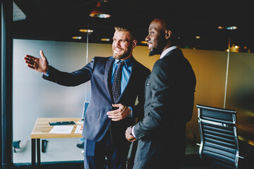 Cheerful caucasian male broker showing office interior African American entrepreneur dressed in formal wear during friendly meeting.Two positive diverse businessmen discussing building planning