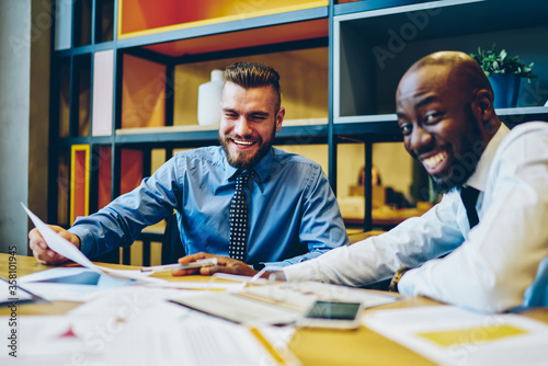 Fotografía Cheerful multiracial male employees in formal wear joking and having fun during working process in office, prosperous skilled economists overjoyed with accomplished report for business plan together