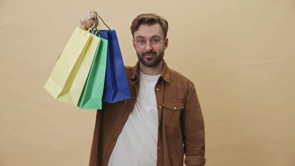 Canvas Print - A charming young unshaven man is showing his colorful purchases standing isolated over beige wall background in studio