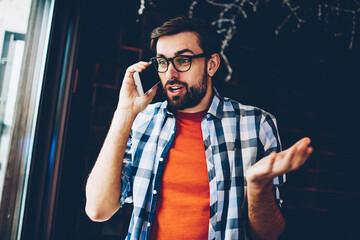 Wall Mural - Amazed bearded young man surprised with news talking with friend during mobile conversation on digital smartphone device.Wondered hipster guy excited with shocked story calling on modern telephone