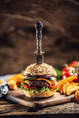 Canvas Print - Knife stabbed in a hamburger with potato wedges on a side in a dark rustic environment