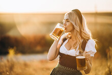 Pretty Happy Blonde In Dirndl, Traditional Festival Dress, Holding Two Mugs Of Beer Outdoors In The Field
