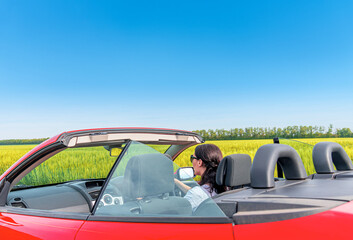 Wall Mural - Woman in a red car against the background of a field and blue sky outside the city.