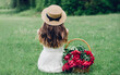 girl in a white dress in a boater sits with her back on the green grass near the basket with red peonies