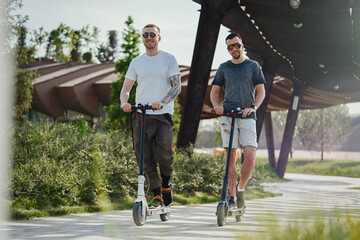 Two handsome men riding electric kick scooters at beautiful park landscape