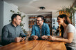 © bnenin - Three smiling coworkers sitting at wooden table, talking and drinking coffee.
