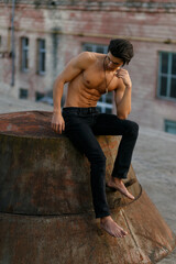 Vertical view of a handsome shirtless, sexy brunette young man with tanned athletic body, seated in profile on old roof at the sunset, looking down.