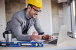 © Dusan Petkovic - Young smiling architect in suit with helmet on head holding smart phone and using laptop while leaning on table in building in construction process.