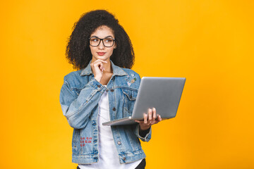 Young african american black positive cool lady with curly hair using laptop and smiling isolated over yellow background.