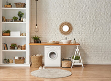 Washing Machine In The Laundry Room, Wooden Table And Shelf Style, Sink Lamp Mirror And Wicker Basket Decor Object.Laundry Room Interior Style, Washing Machine Wicker Basket White Bookshelf And Sink.