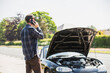 Young man next to his car, depressed and angry on the mobile phone telephone call after the car has broken down on the roadside waiting for assistance to arrive and recover or fix the vehicle