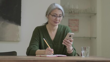 Canvas Print - Calm attractive mature woman with long gray hair in eyeglasses using smartphone while sitting by the table with notebook indoors