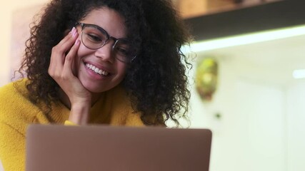 Wall Mural - Cheerful african attractive woman in eyeglasses talking by videocall on laptop computer while sitting on chair indoors