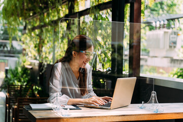 Young 20s Asian beautiful woman wearing a protective face shield and mask with partition in cafe restaurant. While using computer laptop and mobile phone outside. - Corona Virus prevention concept.