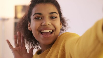 Wall Mural - Close up view of cheerful african attractive woman in yellow sweater posing and making selfie in living room