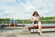 © Roman Tyukin - A woman in a face mask sitting on a bench keeping a social distance with a man sitting on another bench on the promenade. Girl searching for phone in bag and guy using smartphone in social distancing.