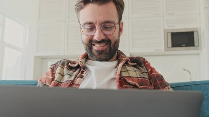 Canvas Print - Handsome young positive cheery bearded man indoors at home sitting on a sofa while using laptop computer