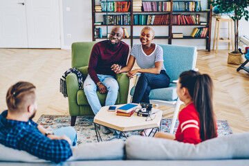 Wall Mural - Smiling african american couple holding hands having conversation with caucasian guests on meeting in apartment, young romantic marriage talking with friends enjoying leisure together at living room