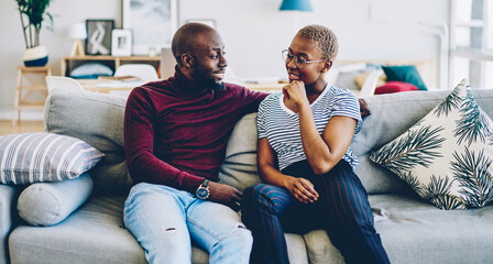 Wall Mural - Young african american marriage  having conversation spending time at home interior together, Handsome dark skinned man looking at his girlfriend hugging sitting on cozy sofa in modern living room.