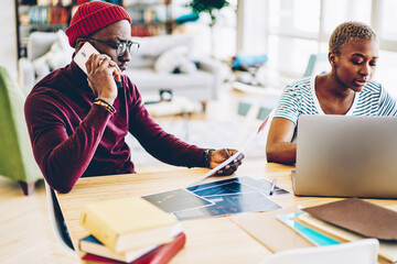 Wall Mural - Serious african american man making telephone call for consultancy with operator while his female colleague using laptop computer for searching information, concentrated coworkers