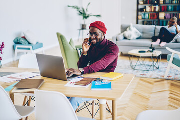 Wall Mural - Happy dark skinned male satisfied with mobile phone conversation with customer support installing app on laptop computer,smiling african american man freelancer satisfied with online business.