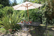 Landscape Of Summer Garden With Outdoor Seating Table And Metal Chairs With Giant Parasol Wood On Stone Patio Outdoors And Exotic Tropical Plants, Bamboo And Hedging, Grass Lawn, Plants In Background