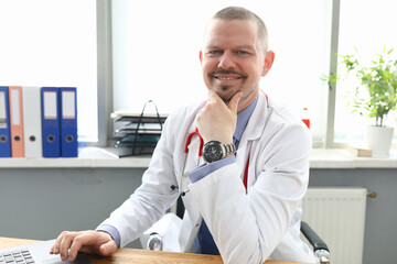 Portrait of smiling professional doctor posing in office. Qualified male wearing uniform and red stethoscope. Laptop on wooden desktop. Medicine and healthcare concept