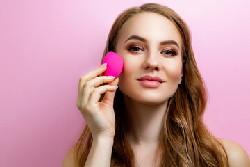 Portrait of a beautiful girl holding a beauty blender for makeup in the studio on a pink background