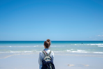 Girl traveling with a backpack stands looking into the distance on a beach with white sand against a background of blue azure transparent water and a blue sky with a clear horizon line.