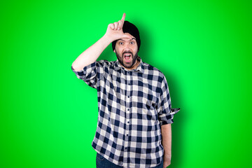 Closeup portrait serious young bearded man showing loser sign on forehead looking at you with disgust at camera isolated green studio background. Negative human emotion facial expression feeling