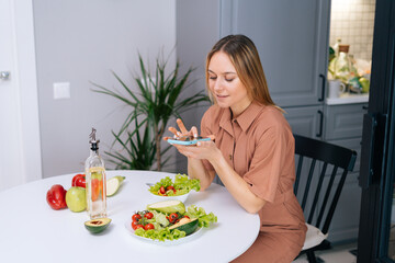 Happy young vegetarian woman uses smartphone for taking photos of vegetables lying on kitchen table. Concept of social networks. Female taking photos of vegetarian food on cell smart phone.