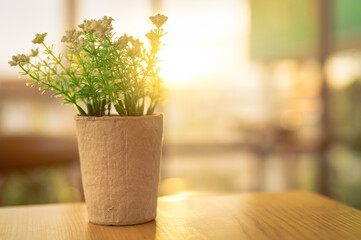 Fake white flowers with green leaves in recycled paper pot on brown wooden table in coffee cafe in the morning with sunshine. Plastic flower in pot. Home office interior and living room decor concept.