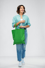 people concept - portrait of happy smiling young woman in turquoise shirt and jeans walking with green reusable canvas bag for food shopping on grey background