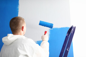 Close-up of professional male worker painting wall with blue paint using roller for work. Change interior design in new apartment. Metal ladder. Renovation and construction site concept