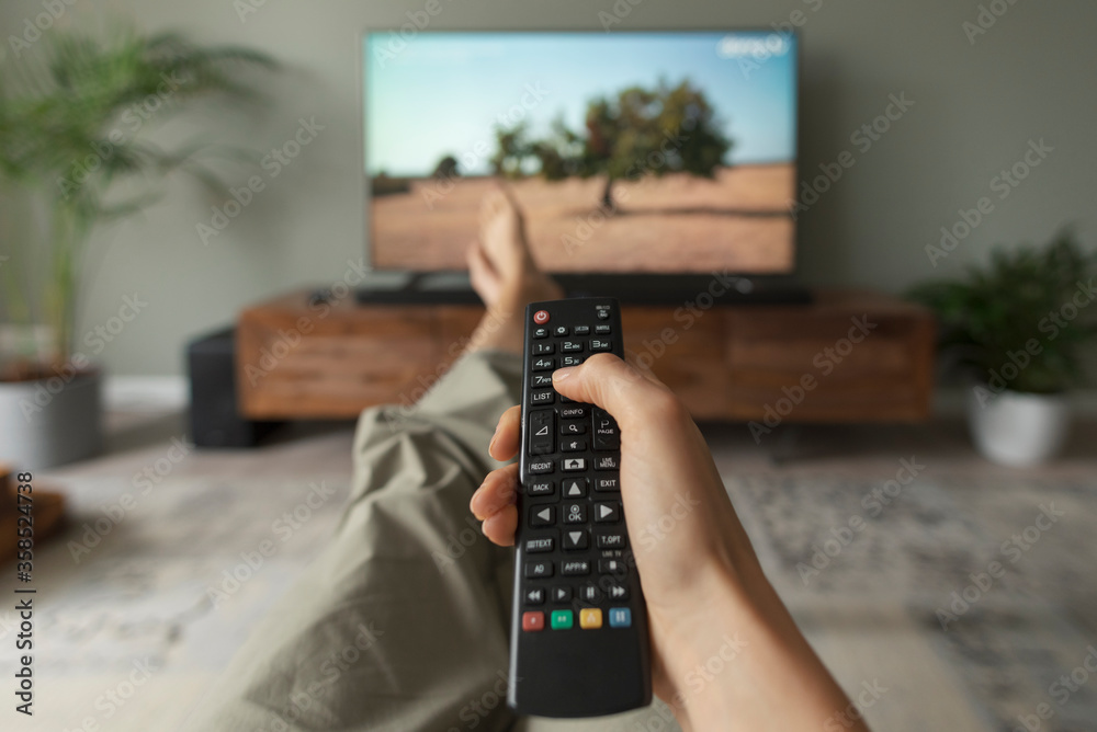 Woman watching TV at home and holding the remote control - obrazy, fototapety, plakaty 