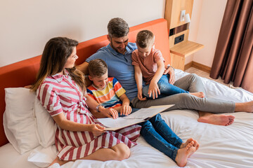 Wall Mural - family of four enjoying together at hotel room, sitting at bed reading hotels offers.