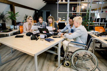 Group of five young creative multiethnic business people working together in modern office with male colleague in a wheelchair for inclusion. Successful disabled business people