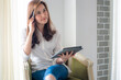 © themorningglory - Happy asian businesswoman using digital tablet sitting on sofa in bedroom over curtain background