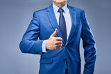 Fototapeta Pomosty - A businessman in a blue suit on a gray-blue background holds his hand in front of him in a sign of approval. The concept of making the right choice and approving the decision.